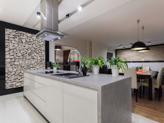 Modern kitchen with a gray stone countertop, stainless steel range hood, and stone accent wall, decorated with potted plants. Dining area with pendant lighting in the background.