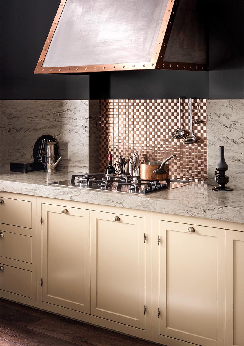 Traditional kitchen design featuring cream cabinetry with brass handles, a marbled countertop, and a copper backsplash with a checkered pattern.