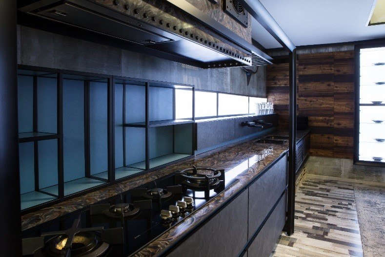 Industrial-style kitchen with dark tones, polished stone countertop, built-in gas stovetop, open black metal shelving, and wood-paneled accents.