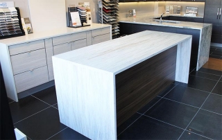 Modern kitchen featuring a large white island with a wooden side panel, gray cabinetry, and a granite countertop in the background.